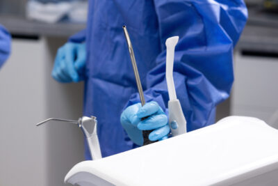 ¿Qué es la cirugía preprotésica y por qué es clave para el éxito de tus rehabilitaciones? Medical theme, female hands wearing gloves and a surgical gown, holding a saliva ejector, dentistry.