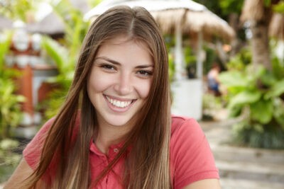 People and lifestyle. Pretty girl with long hair wearing polo shirt smiling happily, relaxing outdoors in exotic country during her vacations, standing against green tropical trees at cafe in open air